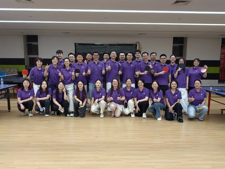 The whole team poses in the table-tennis hall, everyone in the brand purple kit, many holding paddles and giving thumbs-up