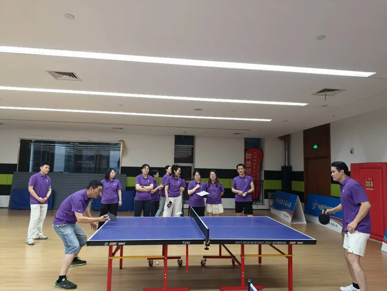 Two colleagues rally across the table, one crouching to receive serve, a row of teammates in the brand purple kit watching courtside